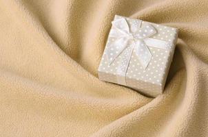 A small gift box in orange with a small bow lies on a blanket of soft and furry light orange fleece fabric with a lot of relief folds. Packing for a gift to your lovely girlfriend photo