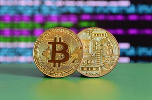 Two gold bitcoins lie on the green surface on the background of the display, which shows the process of mining the crypto currency photo