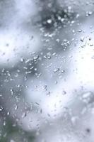 A photo of rain drops on the window glass with a blurred view of the blossoming green trees. Abstract image showing cloudy and rainy weather conditions