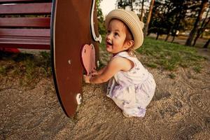 Baby girl in panama hat sat down by the bench at playground outdoor. photo