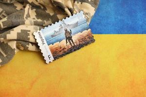 TERNOPIL, UKRAINE - SEPTEMBER 2, 2022 Famous Ukrainian postmark with russian warship and ukrainian soldier as wooden souvenir on army camouflage uniform and national flag photo