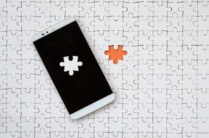 A modern big smartphone with several puzzle elements on the touch screen lies on a white jigsaw puzzle in an assembled state with missing elements photo