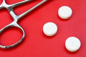 Several white tablets and a medical clamp lie on a bright red background surface. Background image on medical and pharmaceutical topics photo