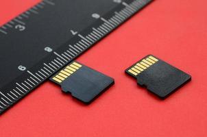 Two small micro SD memory cards lie on a red background next to a black ruler. A small and compact data and information store photo