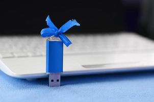 Brilliant blue usb flash memory card with a blue bow lies on a blanket of soft and furry light blue fleece fabric beside to a white laptop. Classic female gift design for a memory card photo