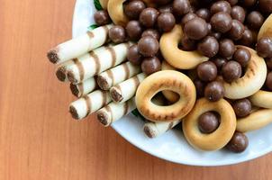 túbulos crujientes, bolas de fusión de chocolate y bagels yacen en un plato blanco sobre una mesa de madera. mezcla de varios dulces foto