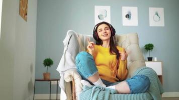 Young girl in headphones with closed eyes relaxes on a cozy sofa and listens to music at home. Attractive joyful woman in a yellow sweater is resting on the couch and singing along in a bright room. video