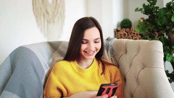 Relaxed Woman in Yellow Sweater Smiling Hold Smartphone Watching Social Media Stories Video Sit on Sofa Home. Happy Girl Look Cellphone Laughing Enjoying Using Mobile App Having Fun Play and Chatting