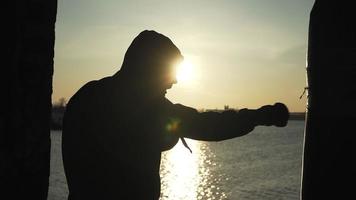 silhouet van een bokser tegen de backdrop van een mooi zonsondergang en een rivier- in een verlaten gebouw, treinen slagen Aan een ponsen tas. episch video. motivatie. sport- en krijgshaftig kunsten. langzaam beweging. video