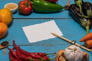 Vegetables are laid out around a sheet of paper and a pencil. Empty space for text. Vegetables, empty blank for recipe on a blue background. photo