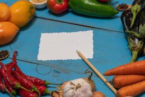 Vegetables are laid out around a sheet of paper and a pencil. Empty space for text. Vegetables, empty blank for recipe on a blue background. photo