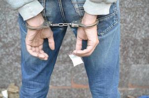 Arrested drug dealer in police handcuffs with small heroin drug package on dark wall background photo