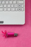 la tarjeta de memoria flash usb de color rosa brillante con un lazo rosa se encuentra sobre una manta de tela suave y peluda de color rosa claro junto a una computadora portátil blanca. diseño clásico de regalo femenino para una tarjeta de memoria foto