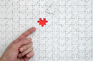 The texture of a white jigsaw puzzle in the assembled state with one missing element, forming a red space, pointed to by the finger of the male hand photo