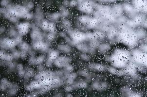 A photo of rain drops on the window glass with a blurred view of the blossoming green trees. Abstract image showing cloudy and rainy weather conditions