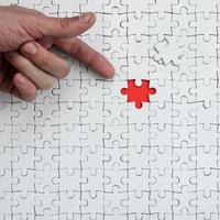 The texture of a white jigsaw puzzle in the assembled state with one missing element, forming a red space, pointed to by the finger of the male hand photo