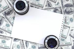 dos lentes fotográficos y un cuaderno blanco se encuentran en el fondo de muchos billetes de dólar. espacio para texto foto