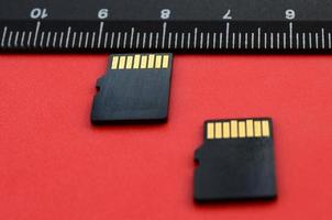 Two small micro SD memory cards lie on a red background next to a black ruler. A small and compact data and information store photo