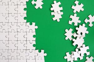 Fragment of a folded white jigsaw puzzle and a pile of uncombed puzzle elements against the background of a green surface. Texture photo with space for text