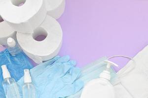 Set of important items for Covid-19 quarantine. Toilet paper, rubber disposable gloves with surgical face mask and hand sanitizer with liquid soap bottle on lilac background photo