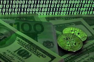 Two bitcoins lies on a pile of dollar bills on the background of a monitor depicting a binary code of bright green zeros and one units on a black background. Low key lighting photo