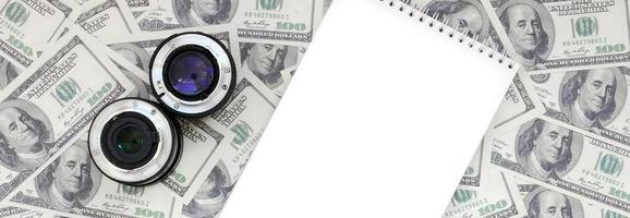 Two photographic lenses and white notebook lie on the background of a lot of dollar bills. Space for text photo