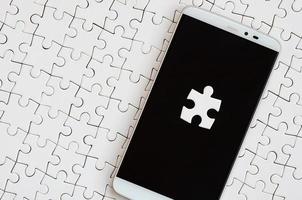 A modern big smartphone with several puzzle elements on the touch screen lies on a white jigsaw puzzle in an assembled state photo