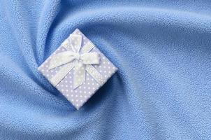 A small gift box in blue with a small bow lies on a blanket of soft and furry light blue fleece fabric with a lot of relief folds. Packing for a gift to your lovely girlfriend photo
