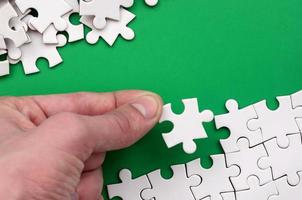 The hand folds a white jigsaw puzzle and a pile of uncombed puzzle pieces lies against the background of the green surface. Texture photo with space for text