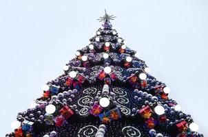 A fragment of a huge Christmas tree with many ornaments, gift boxes and luminous lamps. Photo of a decorated Christmas tree close-up with copy space