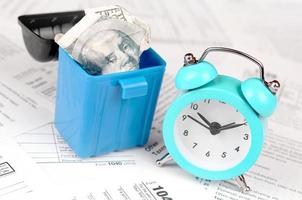 Many american tax blank forms with blue alarm clock and crumpled hundred dollar bill in trash bin photo