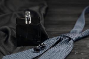 A bottle of mens cologne and cufflinks with blue tie lie on a black luxury fabric background on a wooden table. Mens accessories photo