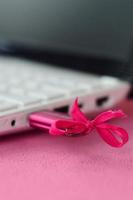 A brilliant pink USB flash drive with a pink bow is connected to a white laptop, which lies on a blanket of soft and fluffy light pink fleece fabric. Classic female design for a memory card photo