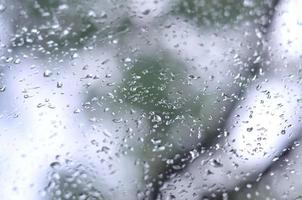 A photo of rain drops on the window glass with a blurred view of the blossoming green trees. Abstract image showing cloudy and rainy weather conditions