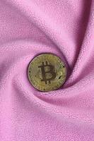 The golden bitcoin lies on a blanket made of soft and fluffy light pink fleece fabric with a large number of relief folds. The shape of the folds resembles a fan from a video card cooler photo