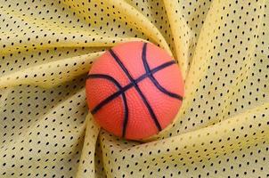 el pequeño baloncesto de goma naranja se encuentra sobre una textura de tela de jersey deportivo amarillo y un fondo con muchos pliegues foto