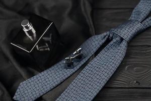 A bottle of mens cologne and cufflinks with blue tie lie on a black luxury fabric background on a wooden table. Mens accessories photo