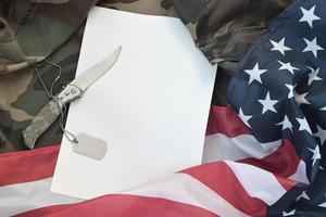 el papel en blanco se encuentra con un cuchillo y un collar de etiqueta de perro del ejército en uniforme de camuflaje y bandera estadounidense foto