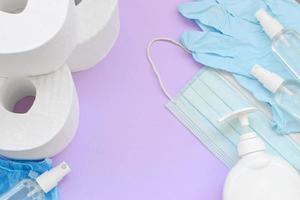 Set of important items for Covid-19 quarantine. Toilet paper, rubber disposable gloves with surgical face mask and hand sanitizer with liquid soap bottle on lilac background photo