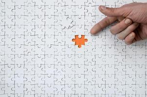 The texture of a white jigsaw puzzle in the assembled state with one missing element, forming an orange space, pointed to by the finger of the male hand photo