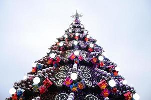 A fragment of a huge Christmas tree with many ornaments, gift boxes and luminous lamps. Photo of a decorated Christmas tree close-up with copy space