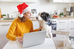 perro y mujer con sombrero de santa con videollamadas familiares por cámara web. chica con laptop usando chat de reunión virtual en vacaciones sentada en la cocina en casa. feliz navidad y año nuevo, nueva normalidad foto