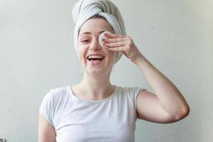 Smiling woman in towel on head with soft healthy skin removing make up with cotton pad isolated on white background photo