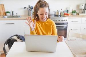 Smiling girl with pet dog waving hand video calling family by webcam. Woman with laptop having virtual meeting chat video call conference on kitchen at home New normal social distance self isolation photo