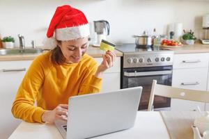 Woman in Santa hat shopping online and paying with gold credit card. Young girl with laptop buying Christmas gift present on Internet on kitchen indoor. Online Christmas shopping concept. photo