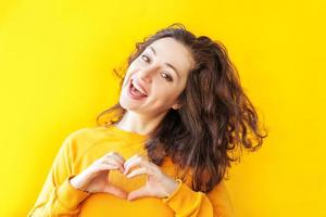 Love, heart shape, peace. Beauty portrait young happy positive woman showing heart sign with hands on yellow background isolated. European girl. Positive human emotion facial expression body language. photo