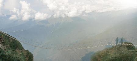 el único camino en las montañas iluminadas por la luz del sol. puente colgante vacío en lo alto de las montañas sobre el fondo de las nubes. foto