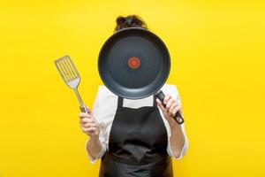 Senior woman cook in a black kitchen apron on a yellow background covered her face with a frying pan, and holds a spatula in her other hand photo