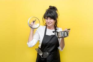 beautiful lady shev chef in an apron with a spatula and a whisk in her pocket holds a pan in one hand, the other lid from it and smiles, yellow background photo
