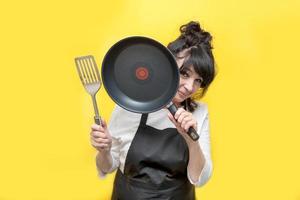 elderly lady cook peeks out from behind a frying pan that she holds in her hand, in the second hand a spatula photo